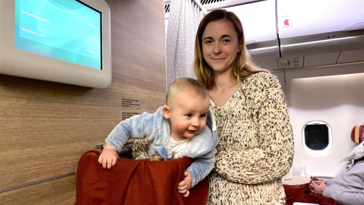Smiling mother standing in an airplane cabin, holding a playful baby next to the airplane bassinet.