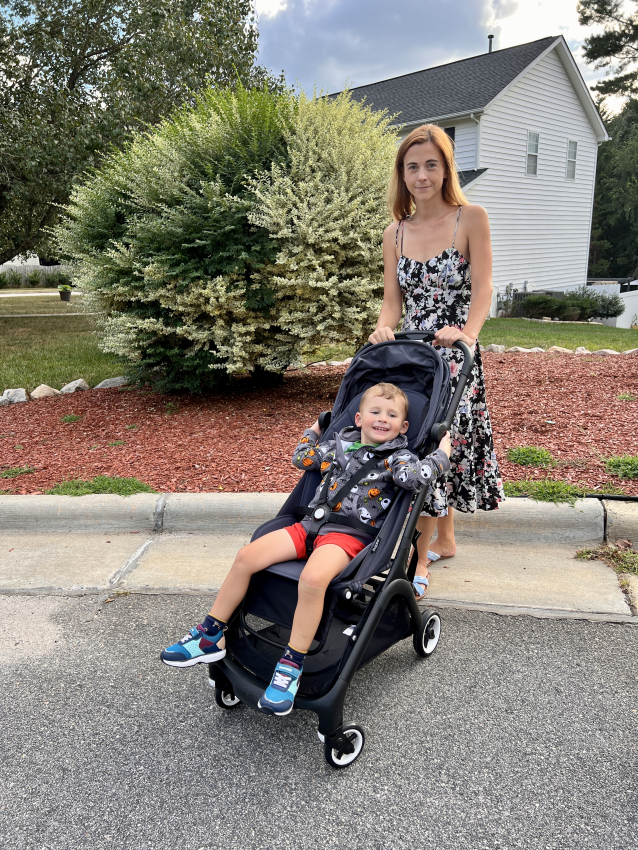 Happy toddler in a Bugaboo stroller with a playful monster print hoodie, accompanied by a woman in a floral dress.
