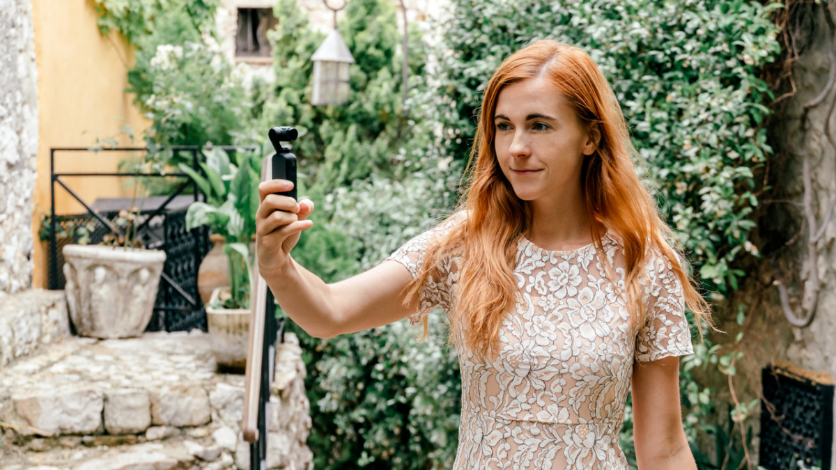 learn language quickly Red-haired woman using a video recorder while exploring a quaint garden setting.