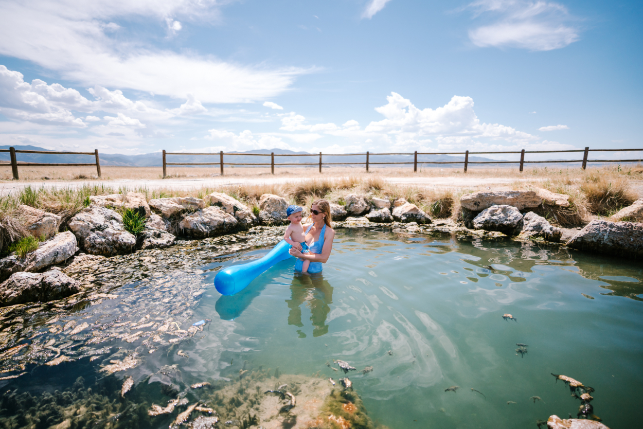 Mother and child enjoy a sunny day in a natural hot spring pool at Meadow Hot Springs with a countryside backdrop, one of Utah's best natural hot springs for families.
