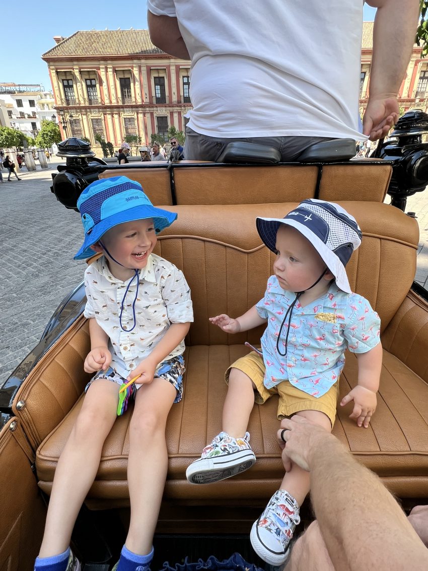 Two toddlers sharing a moment in a carriage ride in a Spanish square, enjoying the sights and sounds of a family-friendly city.